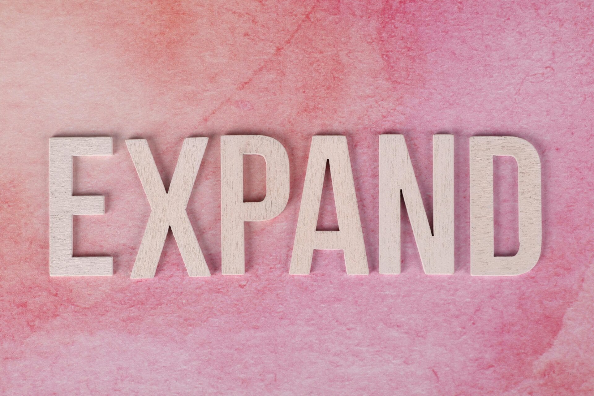Wooden letters spelling 'EXPAND' on a pink textured background, symbolizing growth and creativity.